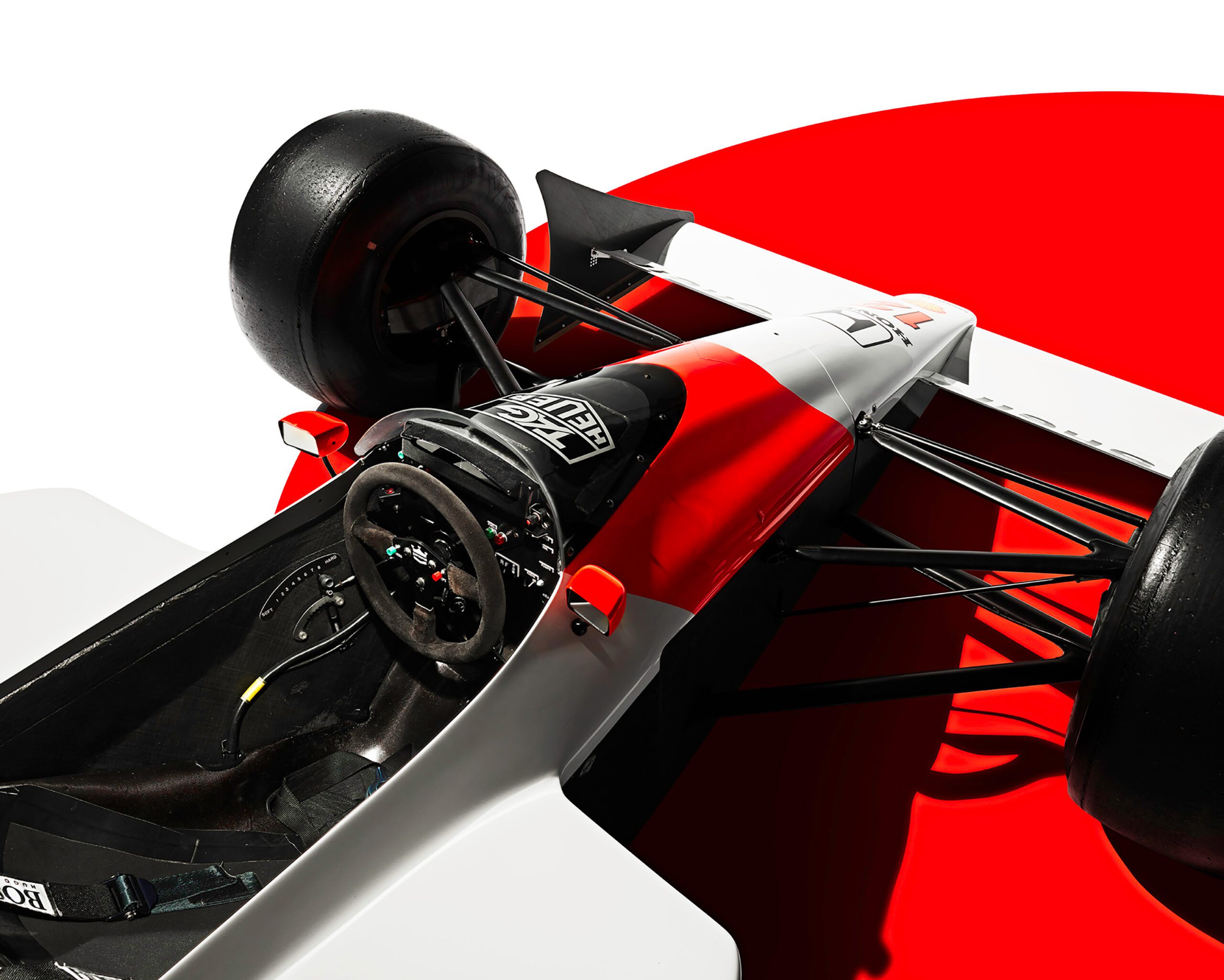 0050_807_WH_McLarenMP44_FrEnd_v3a A top-down view of a Formula 1 race car, reminiscent of Ayrton Senna’s McLaren MP44, featuring a red and white color scheme with black detailing. The cockpit showcases the steering wheel and seat harness. Positioned against a stark white and red background, luxury retouching highlights its sleek design.