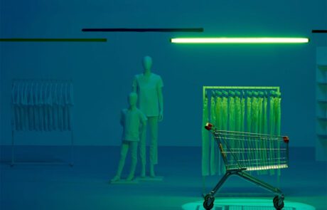 A shopping cart is spotlighted in a dimly lit, blue-tinted store. Mannequins stand near a clothing rack filled with shirts. A green neon light overhead casts an eerie glow on the scene.