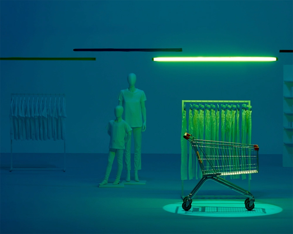 A shopping cart is spotlighted in a dimly lit, blue-tinted store. Mannequins stand near a clothing rack filled with shirts. A green neon light overhead casts an eerie glow on the scene.