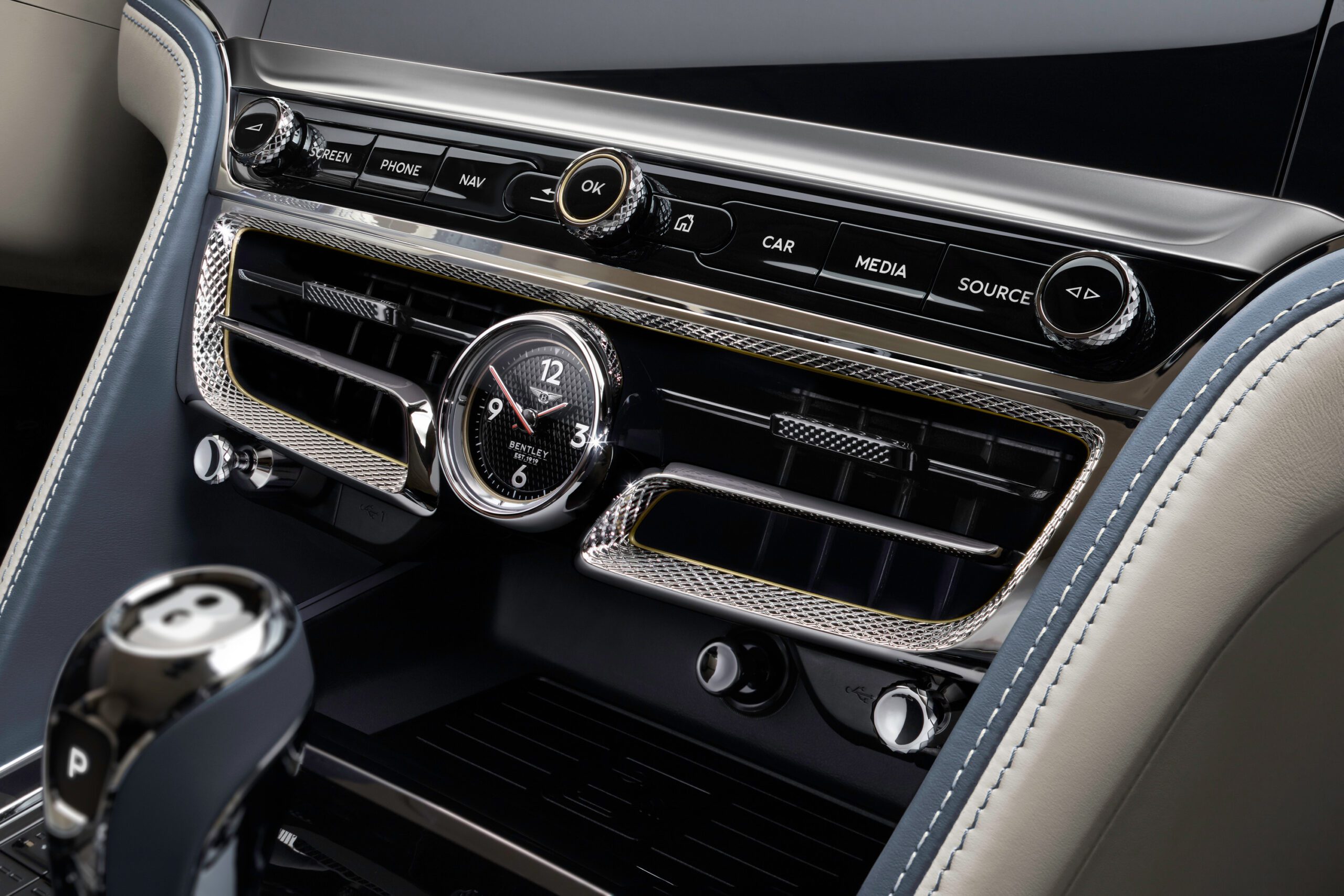 Close-up of the Bentley Flying Spur 2022's dashboard, showcasing a central analog clock with an intricate design, surrounded by various buttons for phone, navigation, media, and car settings. The interior features a combination of metallic and leather textures that any car retoucher would admire.