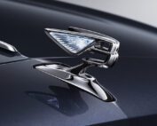 Close-up of a sleek, shiny metallic car hood ornament in the shape of a stylized winged "B" logo, symbolizing luxury. The dark polished surface of the 2022 Bentley Flying Spur reflects the ornament, and the background is blurred to maintain focus on the emblem, showing work worthy of a master car retoucher.