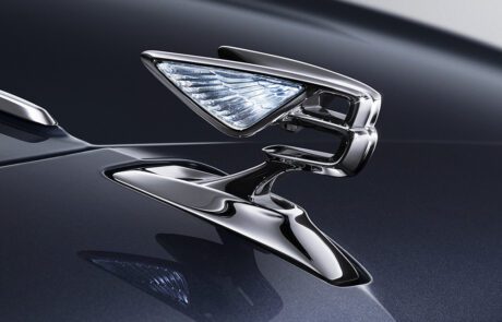 Close-up of a sleek, shiny metallic car hood ornament in the shape of a stylized winged "B" logo, symbolizing luxury. The dark polished surface of the 2022 Bentley Flying Spur reflects the ornament, and the background is blurred to maintain focus on the emblem, showing work worthy of a master car retoucher.