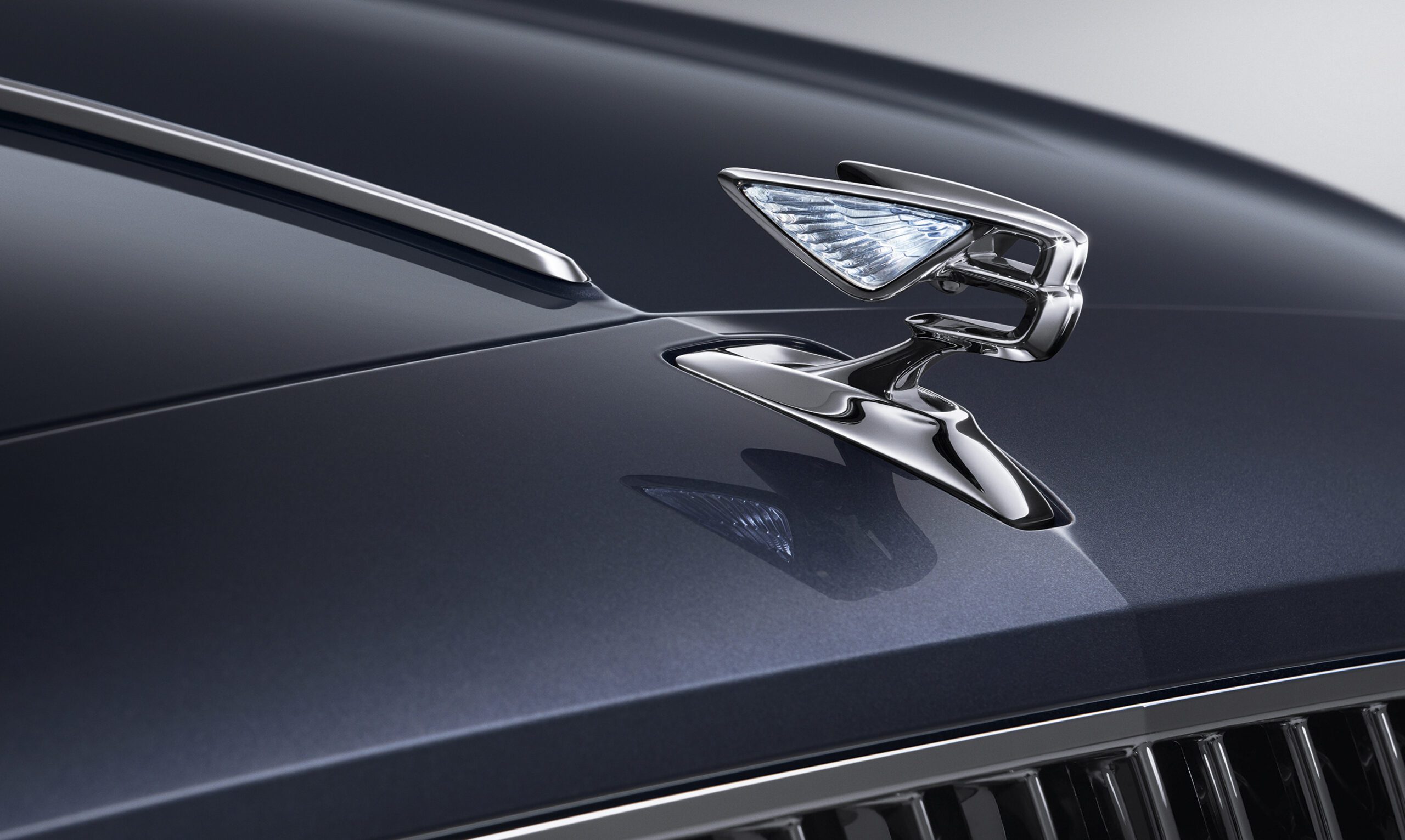 Close-up of a luxury Bentley Flying Spur 2022 hood ornament featuring a sleek, modern design with an illuminated flying-wing style. Meticulously captured by a car retoucher, the chrome-finished ornament stands out elegantly against the smooth, dark paint of the car's hood.