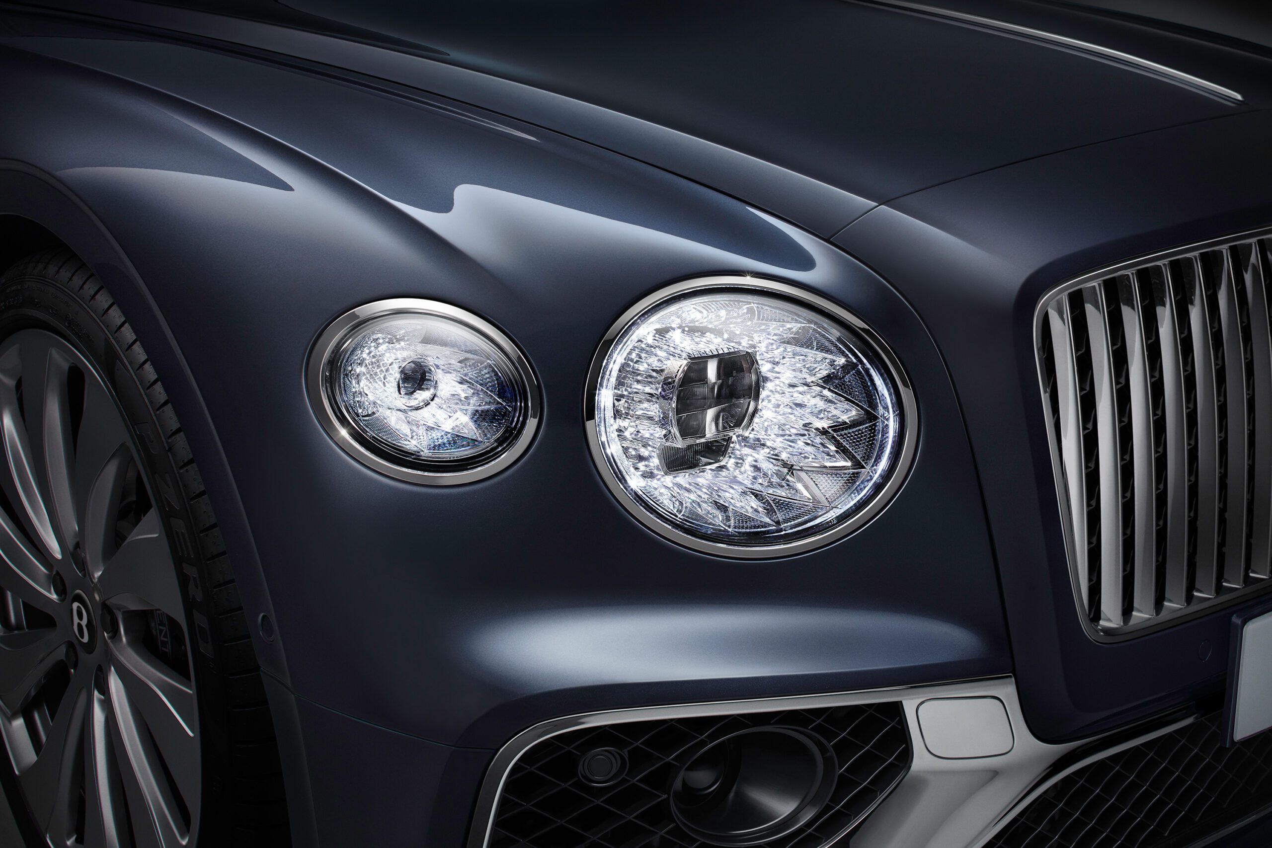 Close-up view of the front right headlight and partial grill of a dark-colored luxury Bentley Flying Spur 2022, showcasing intricate design details and reflections in the headlight, along with part of the front wheel and the iconic car emblem.