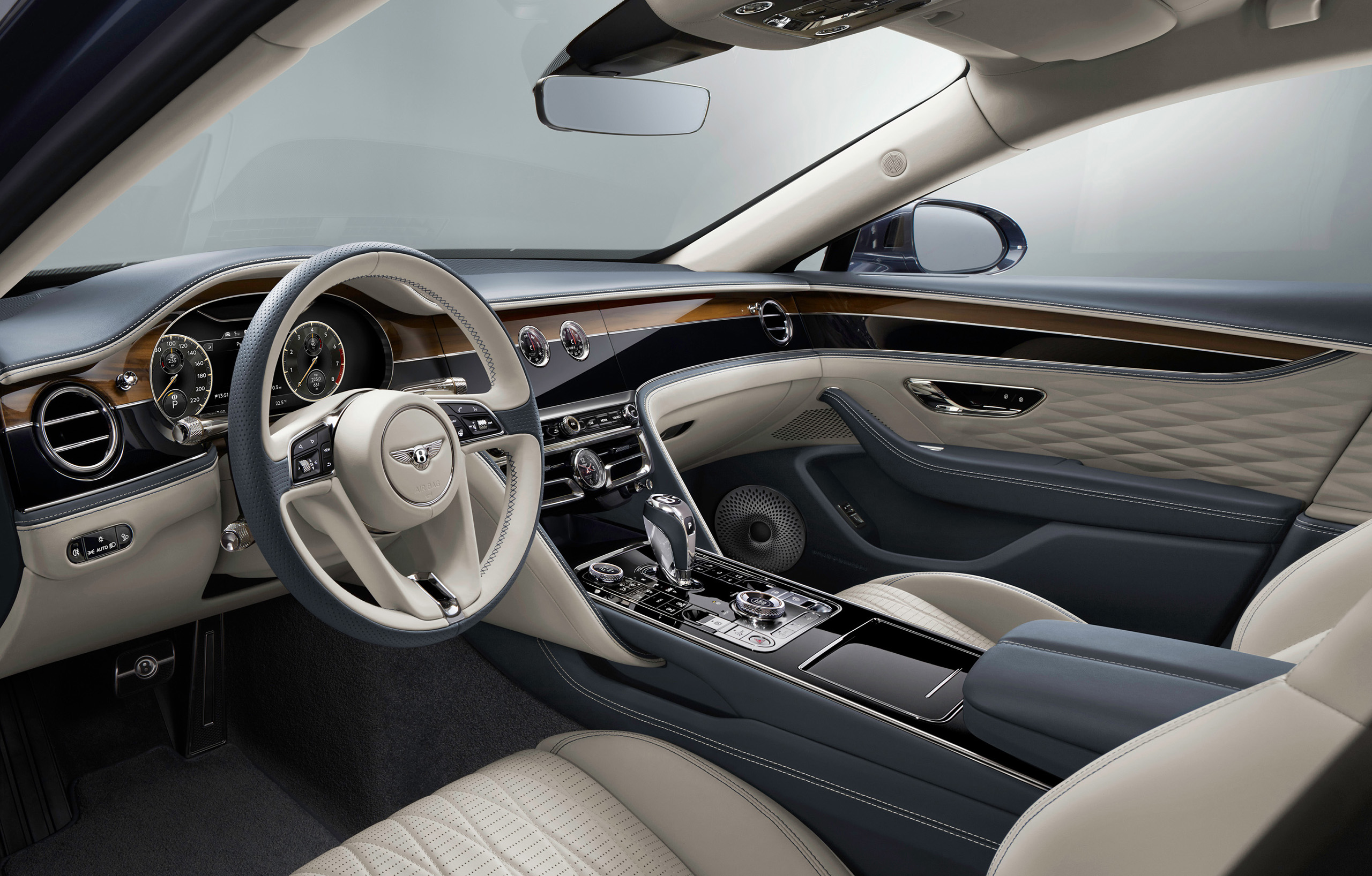 The interior of the Bentley Flying Spur 2022 exudes luxury with a beige and dark blue color scheme. The dashboard has a classic design with analog dials, a touchscreen display, and wooden trim accents. The quilted leather seats are sumptuous, and the steering wheel proudly displays the car brand's emblem.