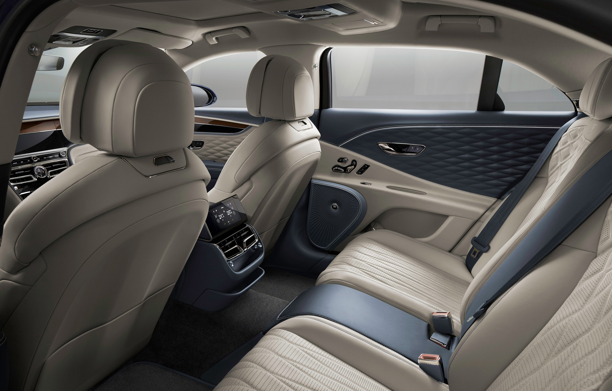 Luxury car interior of the Bentley Flying Spur 2022 featuring beige leather seats with diamond-pattern stitching, spacious legroom, and a high-tech control panel on the rear center console. Wood and metal trim elements adorn the doors and dashboard, highlighting the opulent design.