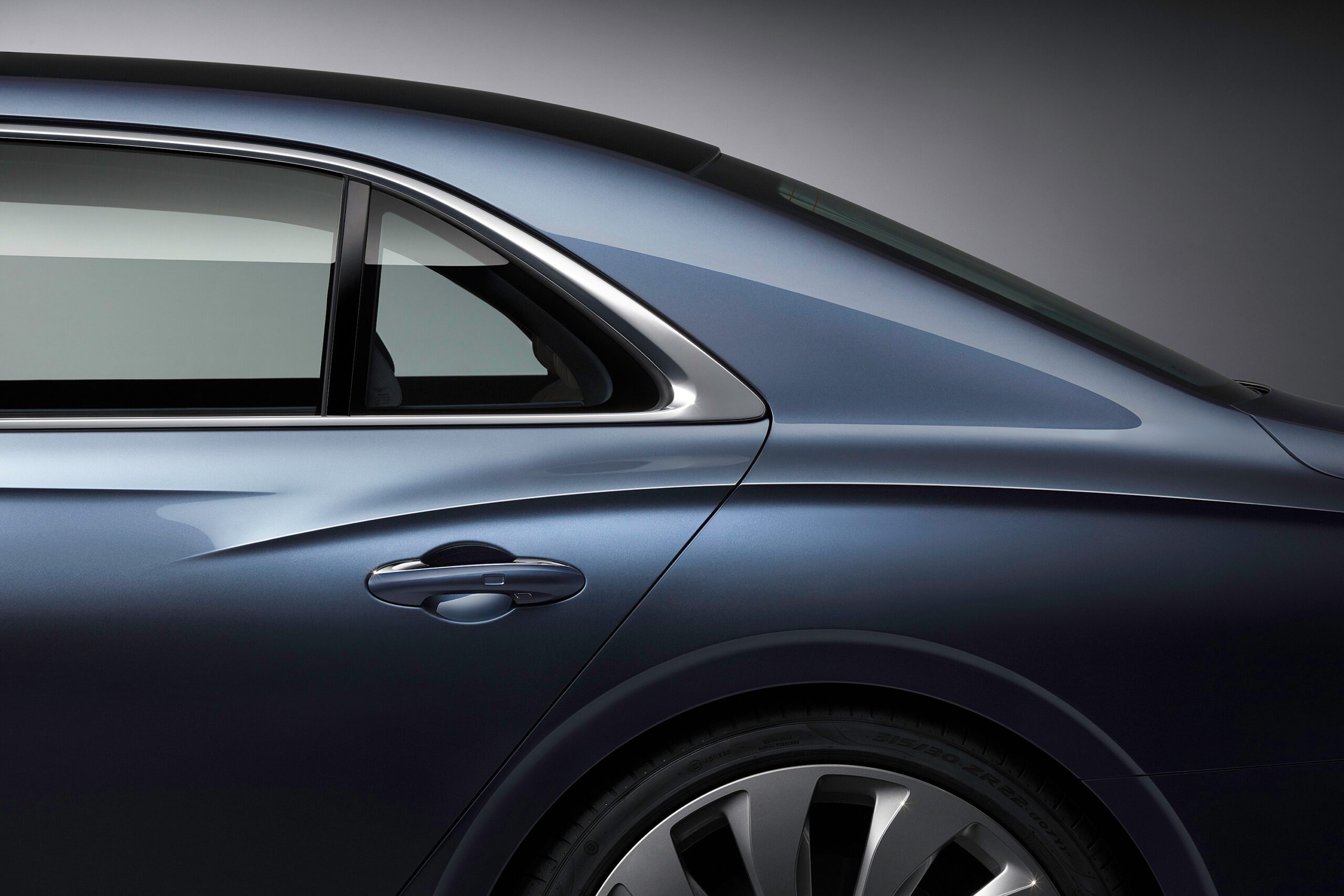 A close-up side view of the rear section of a sleek, dark blue Bentley Flying Spur 2022. The image highlights the smooth lines and contours of the car, including the rear passenger window, door handle, and part of the wheel. A car retoucher's expertise ensures every detail is impeccably showcased against a gradient gray background.