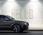 A sleek black Bentley Bentayga Black Edition luxury SUV moves swiftly against a blurred backdrop of a white brick wall with the word "HELLO" subtly illuminated in large letters. The CGI motion blur emphasizes the vehicle's speed and elegance.