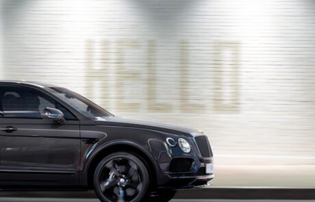 A sleek black Bentley Bentayga Black Edition luxury SUV moves swiftly against a blurred backdrop of a white brick wall with the word "HELLO" subtly illuminated in large letters. The CGI motion blur emphasizes the vehicle's speed and elegance.