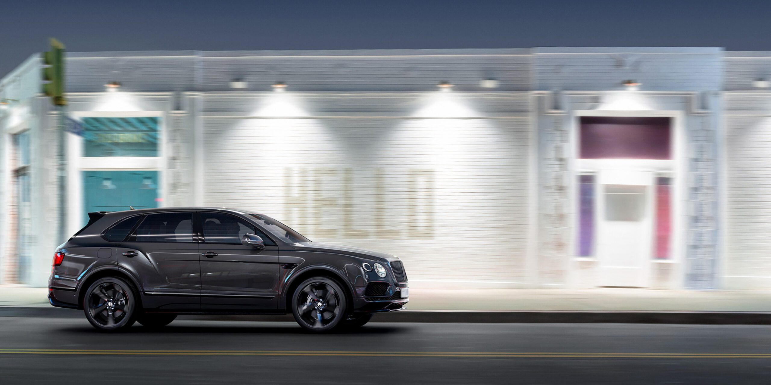 A Bentley Bentayga Black Edition SUV is driving on a city street at night. The background features a well-lit building with a sign that reads "HELLO" in large letters. The scene, possibly crafted with CGI, has a modern, urban atmosphere with a blurred effect indicating motion.