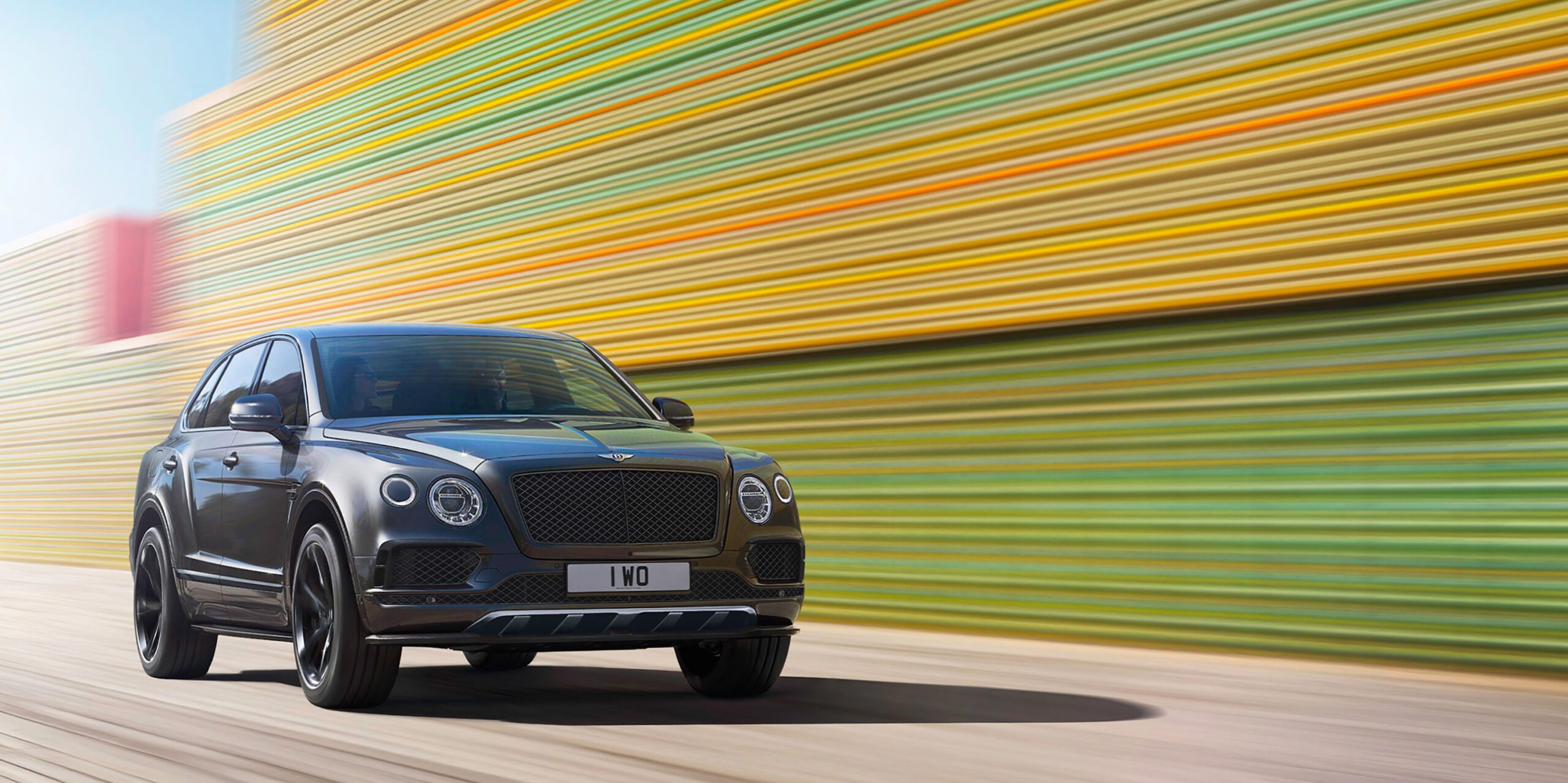 A black Bentley Bentayga Black Edition with a "1 WO" license plate is driving on a road. The background is a blur of colorful horizontal striped patterns, indicating high speed. The luxury SUV features prominent headlights and a sleek design, giving the scene a bright and dynamic, almost CGI-like vibe.