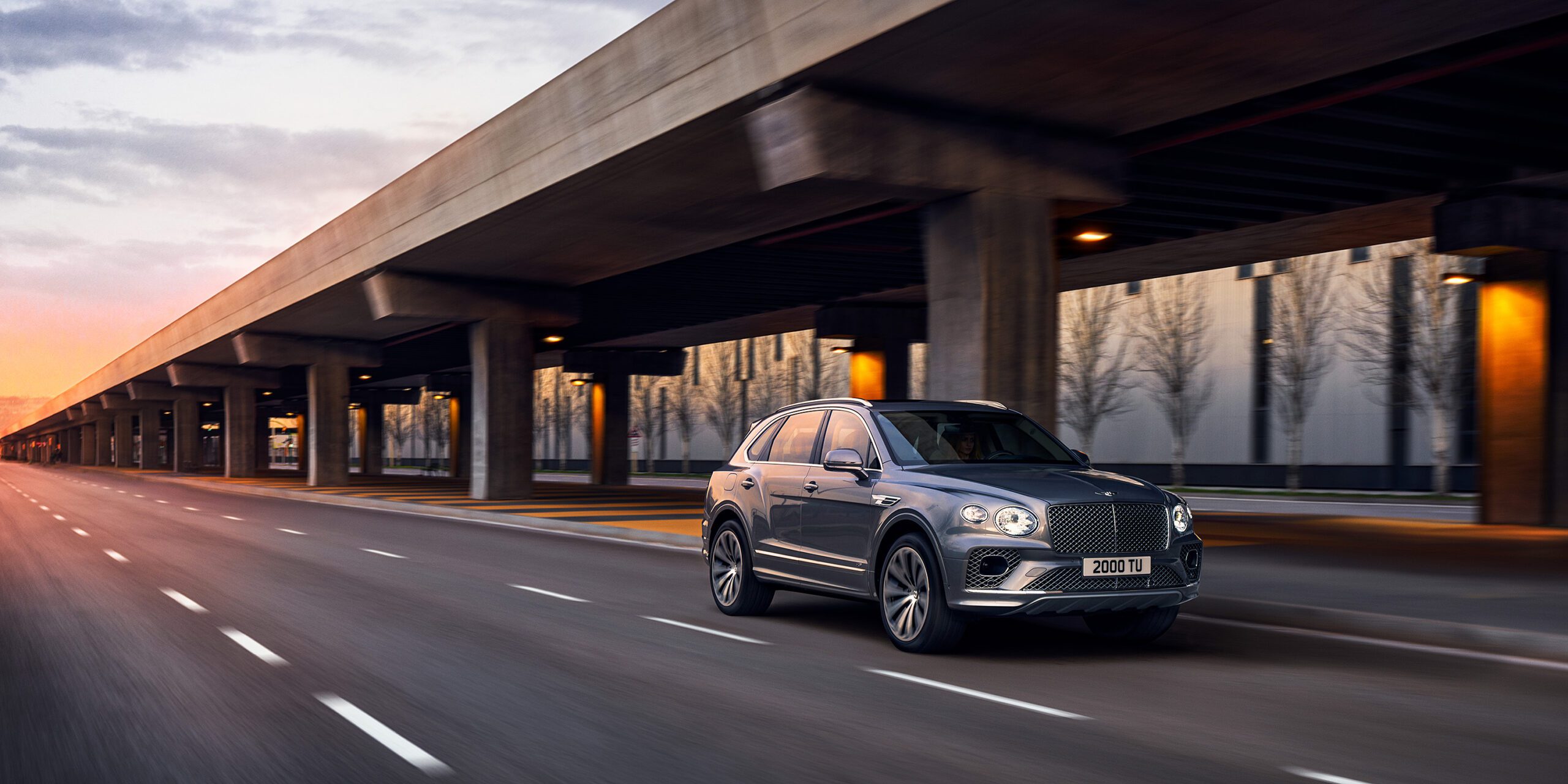 A sleek, blue Bentley Bentayga drives on an empty, multi-lane road under an overpass. The background shows a modern urban setting with trees and buildings at sunset, casting a warm glow on the scene. The vehicle is captured in motion by a high end retoucher London, emphasizing its dynamic design.