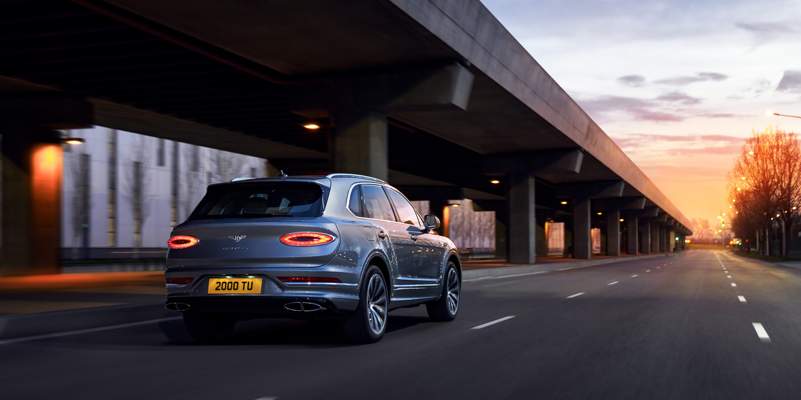 A blue Bentley Bentayga drives along an empty city street at dawn. The vehicle has its taillights on, and the surroundings feature a modern overpass and soft, early morning light with a mix of orange and blue hues. The road is clear, and the sky has a serene, tranquil ambiance.