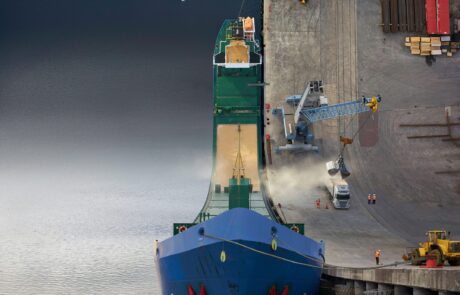 A large blue warped cargo ship is docked at a bustling port in Santander, where loading operations are in progress with a crane, heavy machinery, and stacked containers visible. A small boat navigates the warped world of the busy harbor while a graceful white swan swims serenely near the ship.