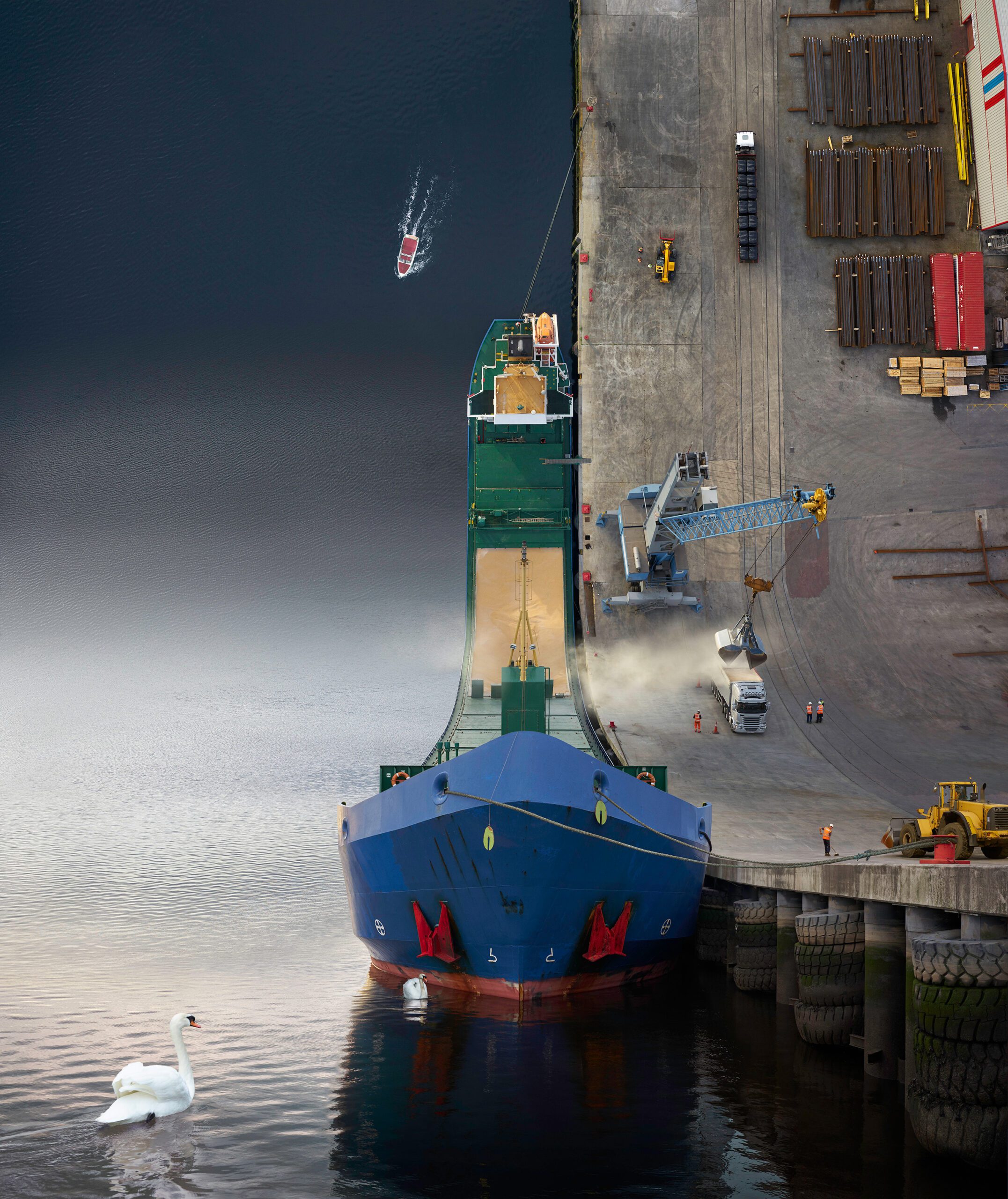 A large blue warped cargo ship is docked at a bustling port in Santander, where loading operations are in progress with a crane, heavy machinery, and stacked containers visible. A small boat navigates the warped world of the busy harbor while a graceful white swan swims serenely near the ship.