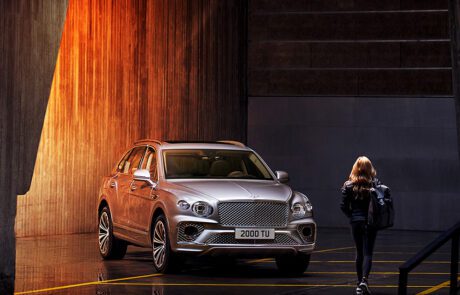 A woman with long blonde hair, dressed in black and carrying a bag, walks towards a Bentley Bentayga parked in an industrial-looking space with dramatic lighting that casts orange hues on the concrete walls. The SUV's license plate reads "2000 TU." A high-end retoucher from London captured this striking scene.