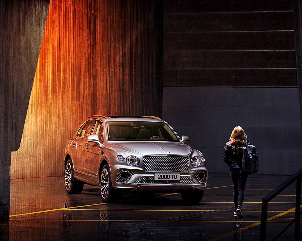 A woman with long blonde hair, dressed in black and carrying a bag, walks towards a Bentley Bentayga parked in an industrial-looking space with dramatic lighting that casts orange hues on the concrete walls. The SUV's license plate reads "2000 TU." A high-end retoucher from London captured this striking scene.