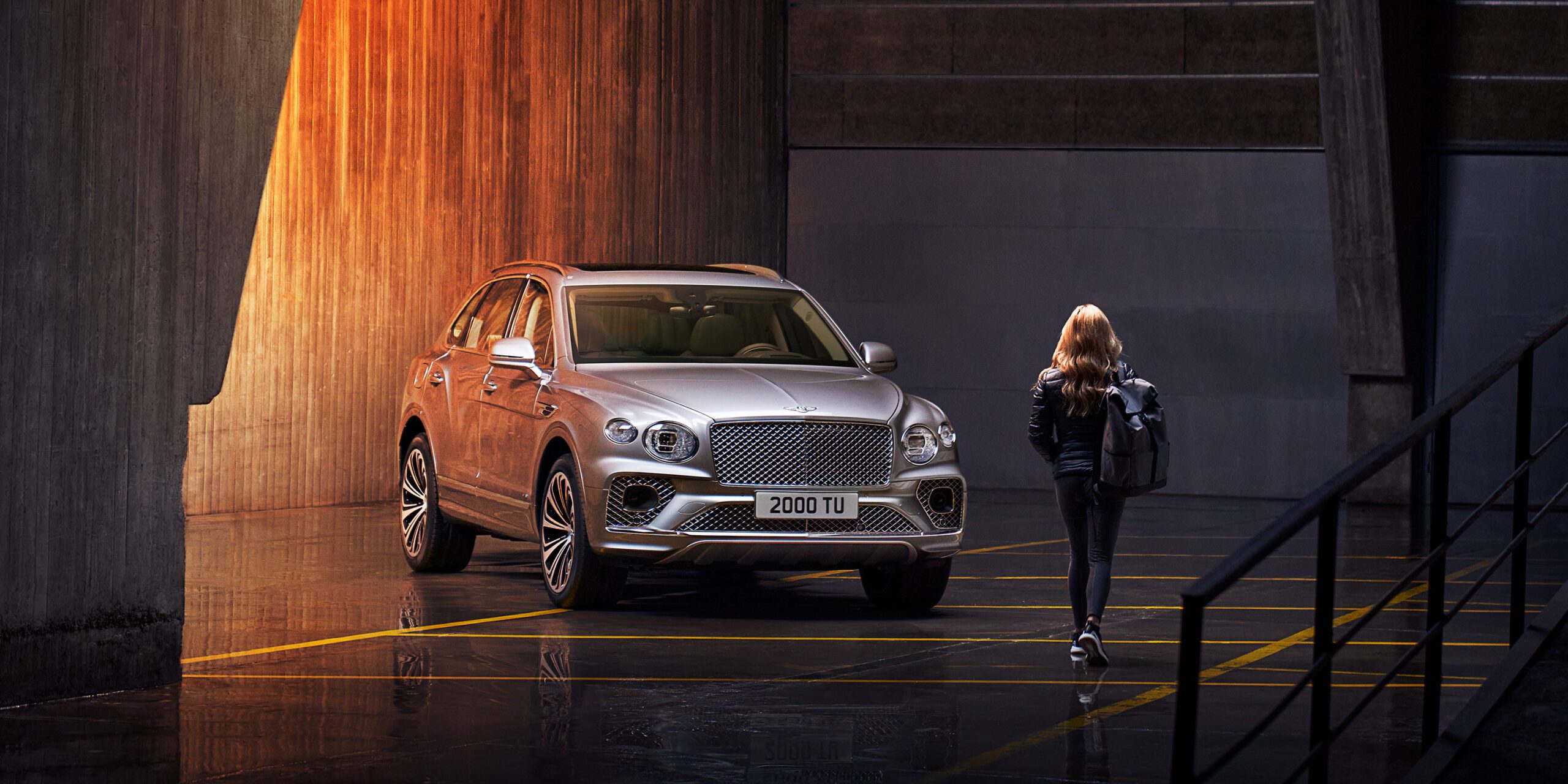 A silver Bentley Bentayga is parked in a dimly lit, industrial-style garage with concrete walls and reflective dark flooring. A person with long hair, wearing a backpack, walks towards the vehicle. The SUV's headlights and details are accentuated by soft lighting, perfectly captured by a high-end retoucher from London.