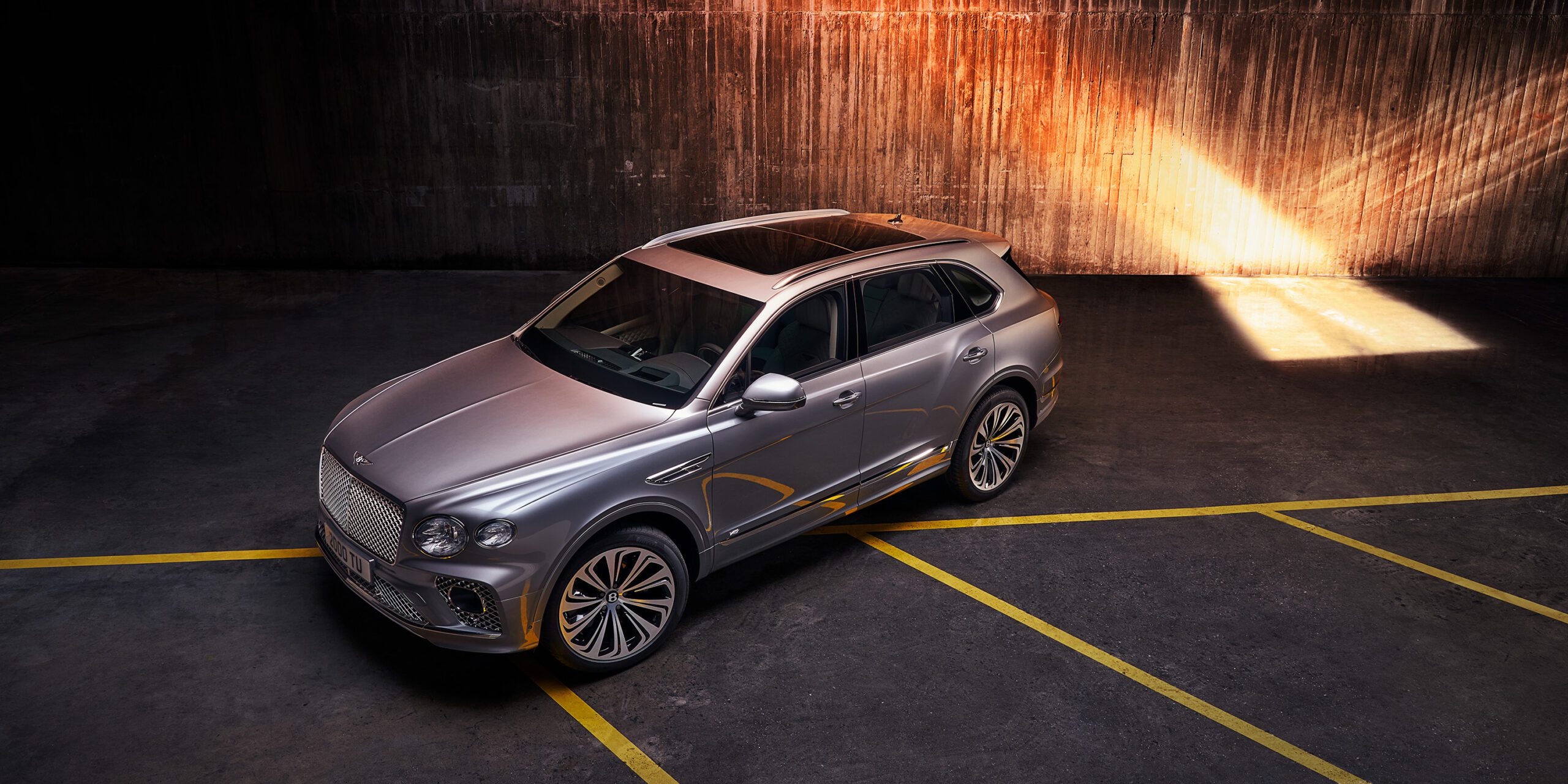 A sleek, silver Bentley Bentayga luxury SUV is parked in an industrial setting, illuminated by a beam of light from above. The setting features a concrete wall casting a shadow, with yellow parking lines accentuating the vehicle's modern and elegant design. Retouched to perfection by a high-end retoucher in London, every detail shines.