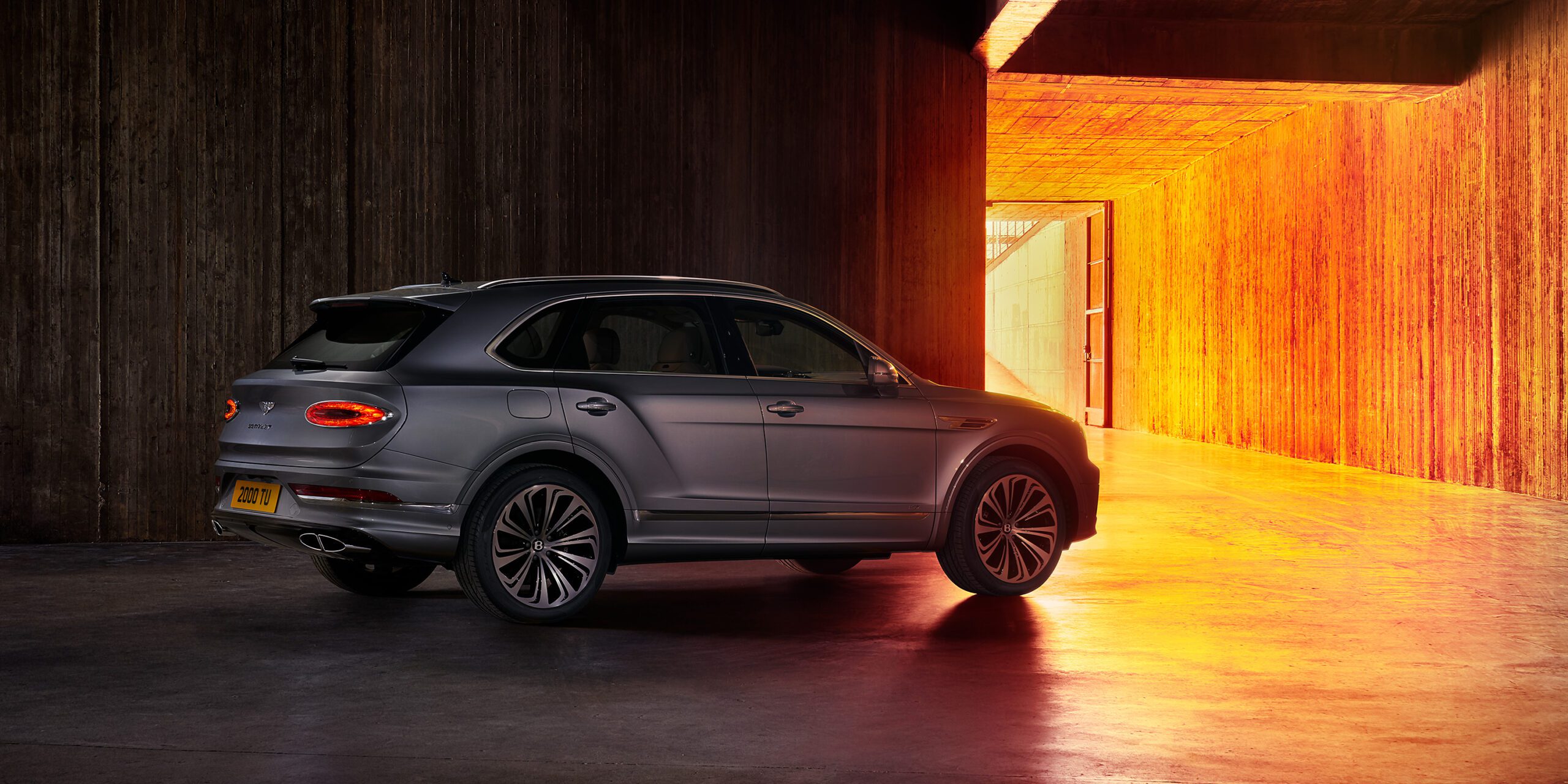 A sleek Bentley Bentayga luxury SUV is parked inside an industrial-style building with a concrete floor and walls. Warm, orange light illuminates the car from a large, open doorway, creating a dramatic contrast against the vehicle's metallic exterior.