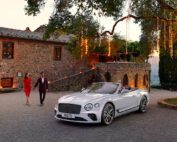 A couple dressed elegantly walks hand-in-hand towards a luxurious white Bentley GT Continental Convertible parked on a gravel driveway. They are in front of a rustic stone building adorned with hanging lights, all showcased flawlessly through expert advertising retouching. The background features a scenic, hilly landscape.