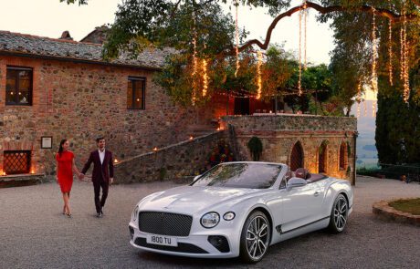 A couple dressed elegantly walks hand-in-hand towards a luxurious white Bentley GT Continental Convertible parked on a gravel driveway. They are in front of a rustic stone building adorned with hanging lights, all showcased flawlessly through expert advertising retouching. The background features a scenic, hilly landscape.