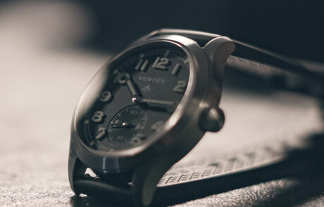 A close-up of a Vertex wristwatch with a black dial, white numerals, and a leather strap, resting on a textured surface in soft lighting.