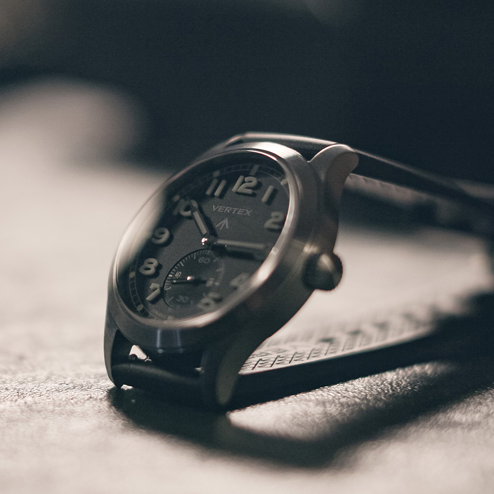 A close-up of a Vertex wristwatch with a black dial, white numerals, and a leather strap, resting on a textured surface in soft lighting.
