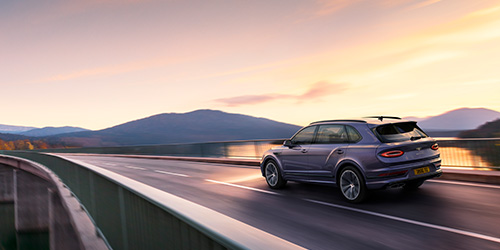 A sleek, dark Extended Wheelbase Bentayga drives across a long, elevated bridge at sunset. The bridge's metal railings frame the road, and distant hills and mountains are silhouetted against a colorful sky. The car's rear is visible, with its taillights and license plate illuminated in this CGI scene.