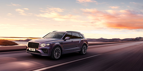 A purple Extended Wheelbase Bentayga luxury SUV is driving on a paved road. The road is empty and stretches towards the horizon. The sky is filled with soft orange and pink hues from the setting or rising sun, with mountains visible in the distance, creating a scene that looks almost CGI-crafted.