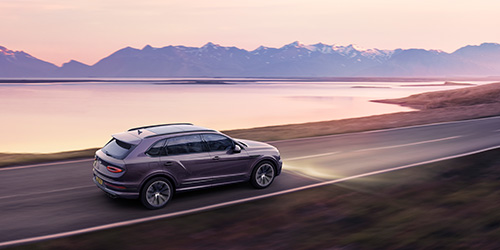 An Extended Wheelbase Bentayga SUV drives along a coastal road at dusk with its headlights on. The surrounding landscape includes a serene body of water, a narrow shore, and distant mountains under a pink and purple sky. It almost looks like something out of CGI.