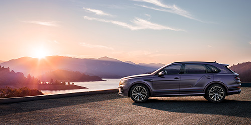 A purple Extended Wheelbase Bentayga is parked on a road overlooking a scenic landscape at sunset. The sun is setting behind a range of mountains with a body of water in the foreground, casting a serene glow on the almost surreal CGI-like scene.