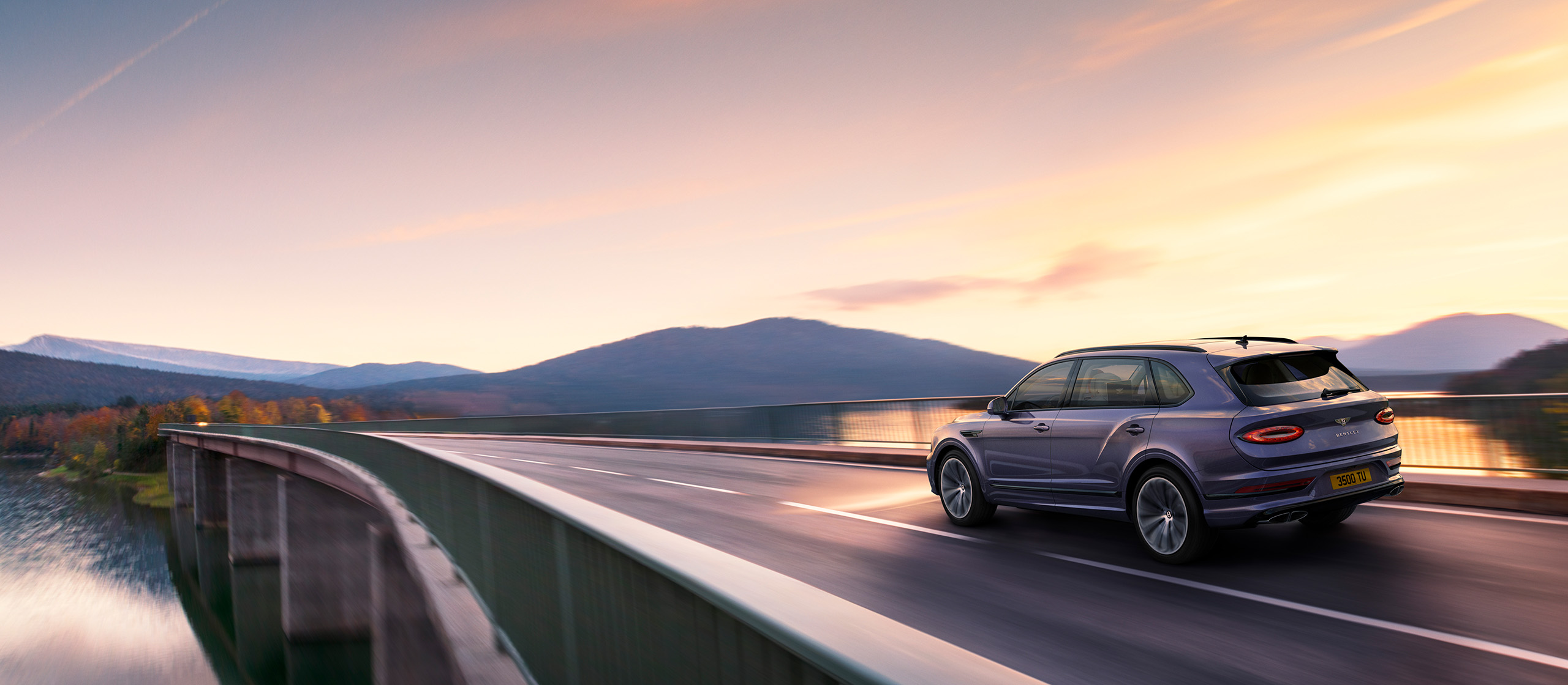 A sleek Bentley Bentayga Extended Wheelbase drives on a winding bridge over a tranquil body of water, with a backdrop of distant mountains and a colorful sunset sky. The scene, rendered in stunning CGI, captures the essence of a peaceful evening drive in a beautiful natural landscape.