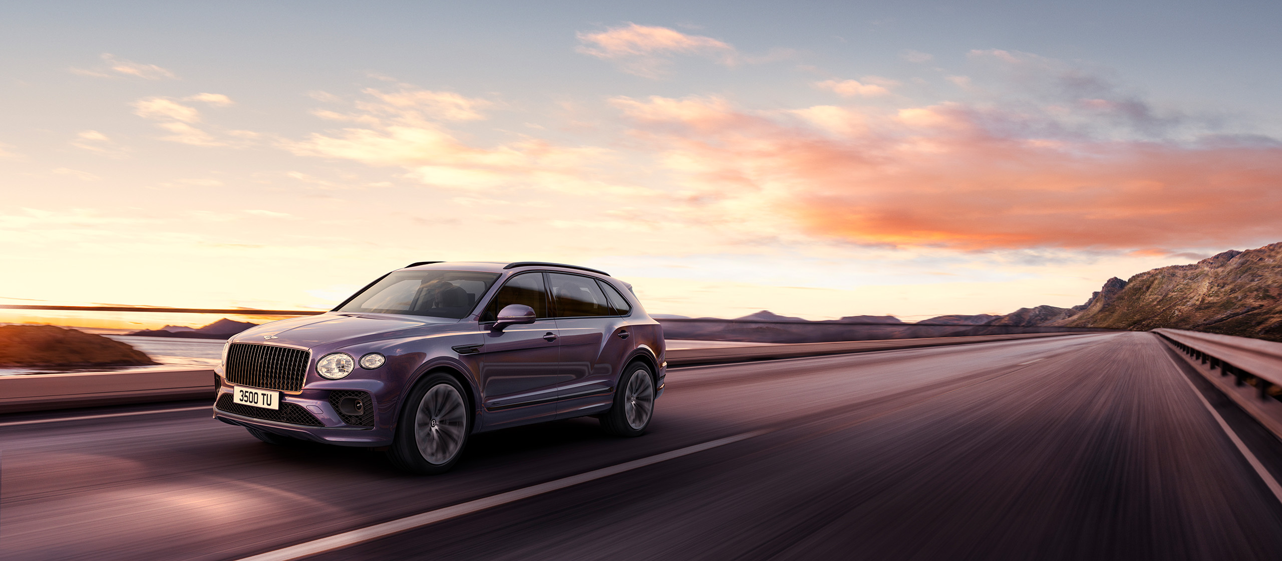 A purple Bentley Bentayga extended wheelbase drives along a coastal highway with mountains in the background. The sky displays a beautiful sunset with hues of orange, pink, and purple. The vehicle moves swiftly on the smooth road, capturing the essence of a serene and scenic journey.