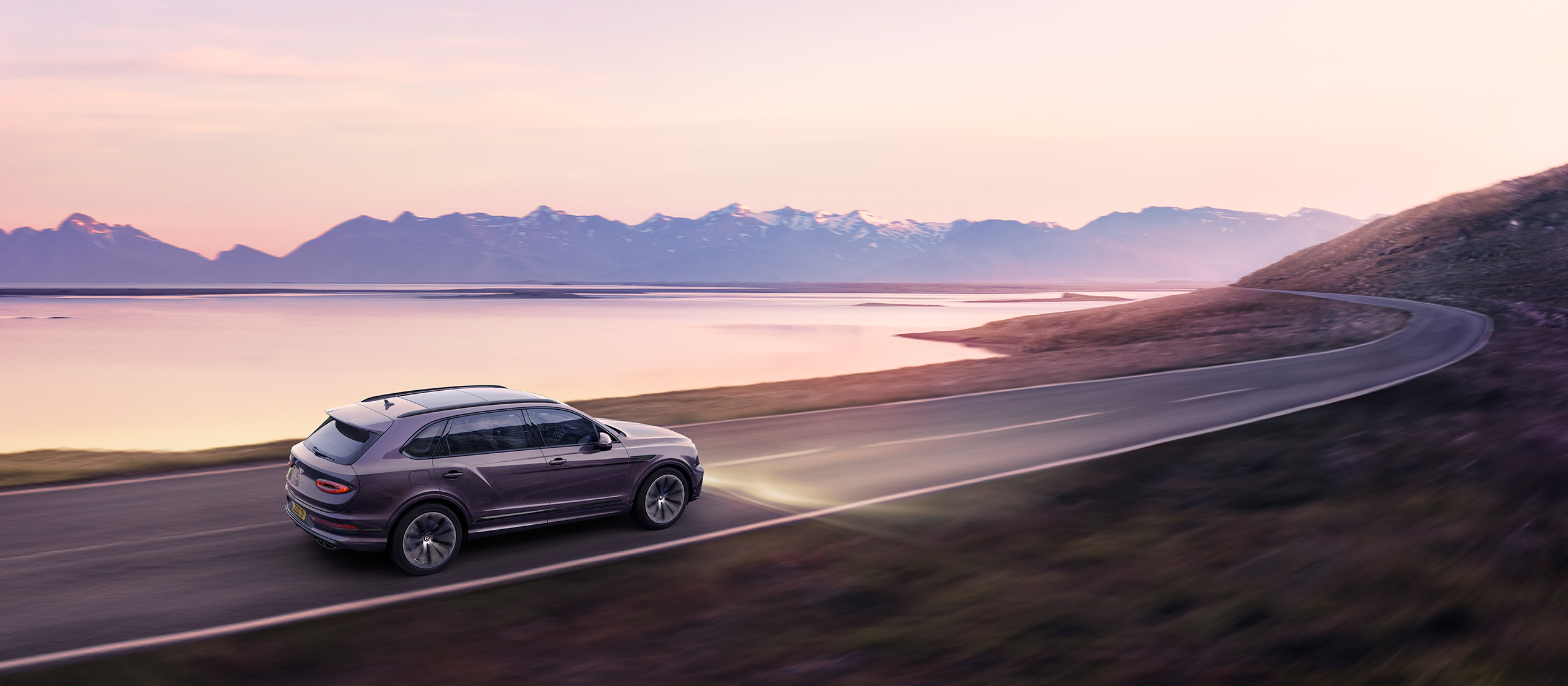 A Bentley Bentayga Extended Wheelbase luxury SUV drives on a scenic, winding road beside a serene lake during sunset. Snow-capped mountains are visible in the background, and the sky is painted in soft shades of pink and purple. The SUV's headlights are on, illuminating the road ahead, almost like a CGI scene.