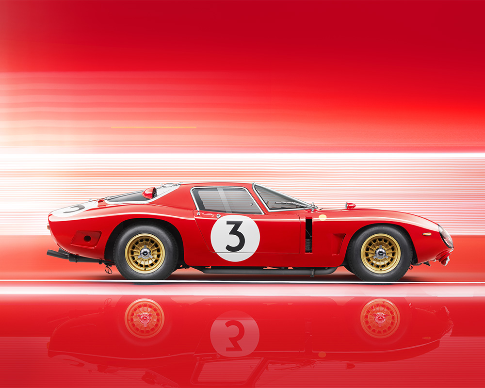 Side view of a sleek, red vintage Bizzarrini 5300 GT Corsa with the number 3 on its door and hood, set against a dynamic red and white blurred background. The car has gold rims and an aerodynamic design, creating a sense of speed and motion in this retouched image.