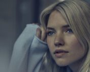 A woman with blonde hair and blue eyes looks thoughtfully to the side, as if contemplating a serene melody. She is wearing a light-colored, long-sleeved top, with one hand resting against her head. The image’s pensive mood feels meticulously crafted.