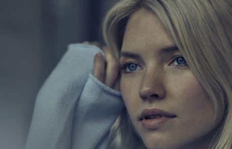 A woman with blonde hair and blue eyes looks thoughtfully to the side, as if contemplating a serene melody. She is wearing a light-colored, long-sleeved top, with one hand resting against her head. The image’s pensive mood feels meticulously crafted by a high-end retoucher.