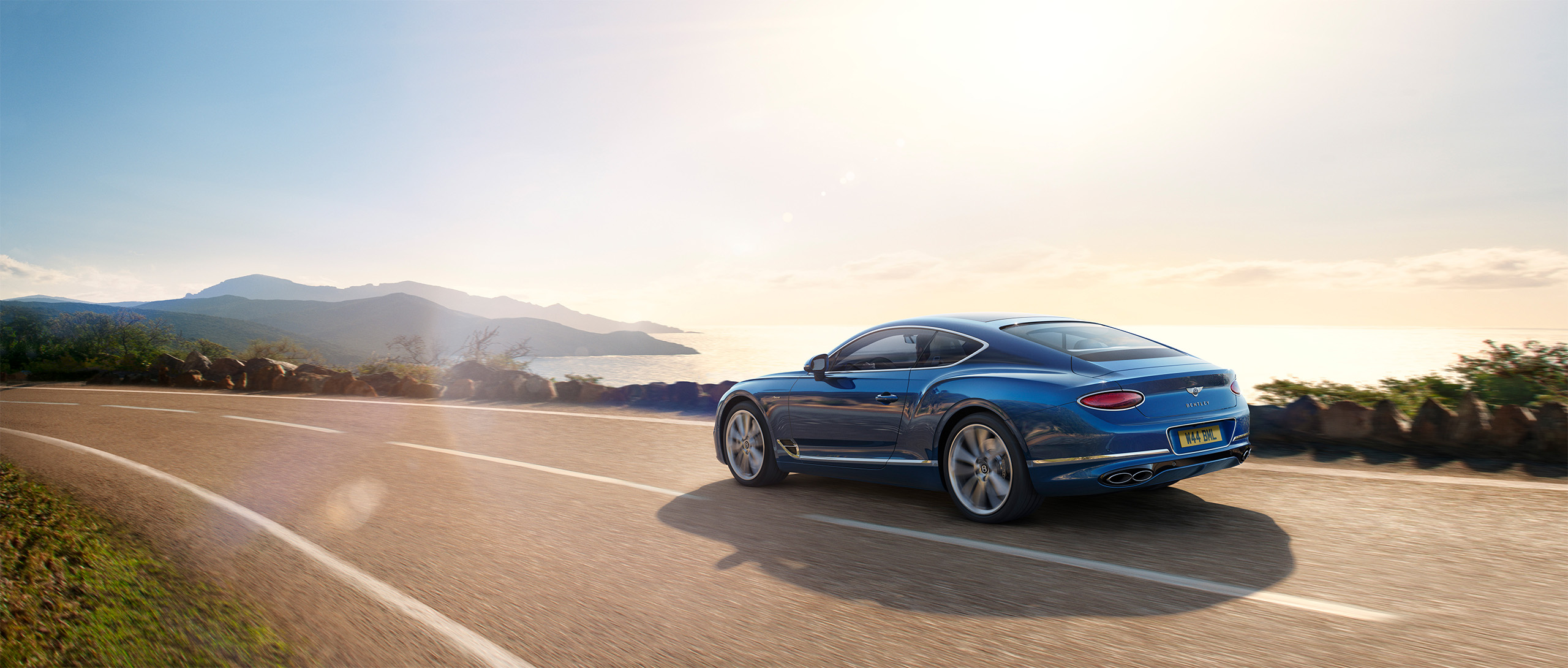 A Bentley Azure from the CGI Studio London drives on a winding coastal road during sunset, with mountains and the ocean visible in the background. The sun casts a warm glow over the scene, and the empty road stretches ahead.