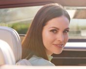 A woman with long brown hair smiles softly while sitting in a Bentley GT Convertible. She is looking over her shoulder towards the camera, with sunlight filtering through the car window. The image captures a warm and serene moment, perfectly highlighted by subtle car retouching.