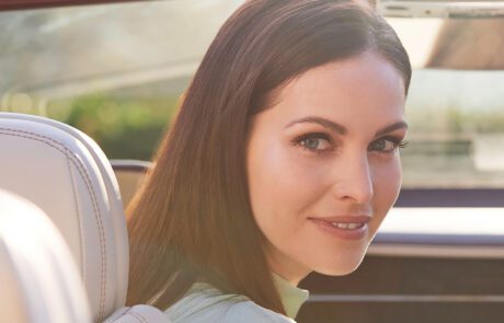 A woman with long brown hair smiles softly while sitting in a Bentley GT Convertible. She is looking over her shoulder towards the camera, with sunlight filtering through the car window. The image captures a warm and serene moment, perfectly highlighted by subtle car retouching.