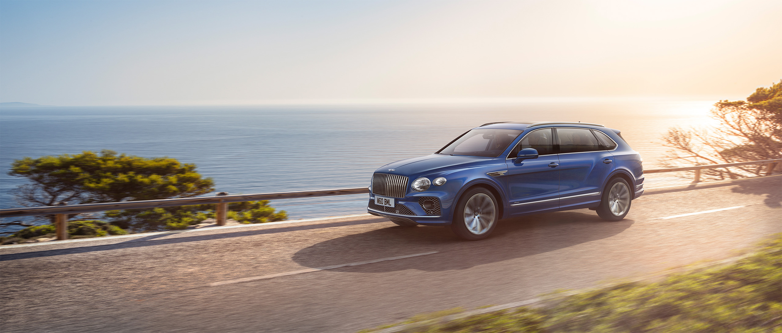 A Bentley Azure SUV is driving on a coastal road with a clear view of the ocean on a sunny day. Trees and bushes line the road, and the sun is setting or rising, casting a warm glow over the scene, as if crafted by a CGI studio in London.