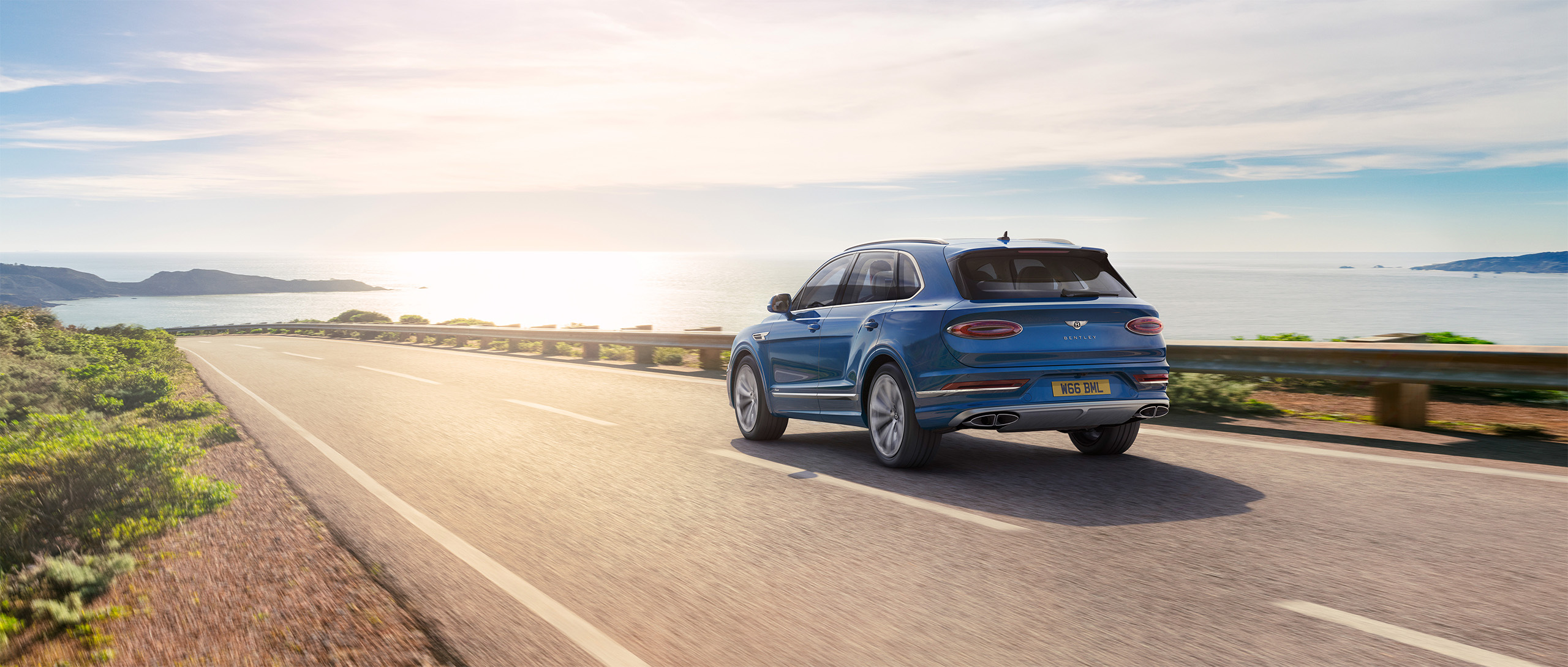 A blue Bentley Azure SUV is driving on an open coastal road with the ocean and cliffs visible in the background under a bright, sunny sky. The sun reflects off the water, creating a serene and picturesque scene of a scenic drive. This breathtaking moment feels like it was crafted by CGI Studio London.