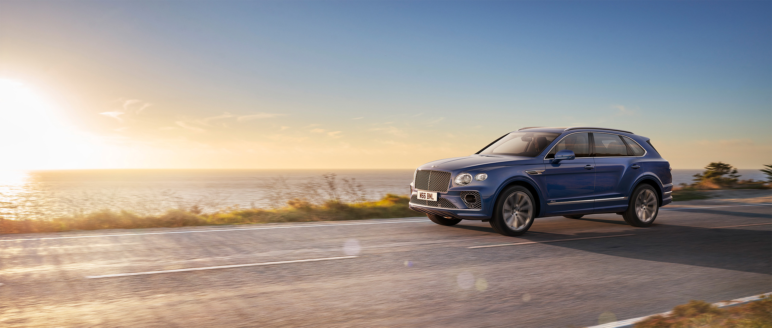 A blue Bentley Azure is driving on a coastal road during sunset. The car is moving away from the camera, with the ocean and the setting sun in the background. The sky displays a gradient of warm colors transitioning into cooler tones as it meets the horizon, captured flawlessly by CGI Studio London.