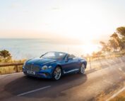 A sleek blue Bentley Azure convertible car is driving on a coastal road at sunset. The sea and distant shoreline are visible in the background, with the sun casting a golden glow over the scenery. Tall grass and trees partially line the road, as if crafted by CGI Studio London.
