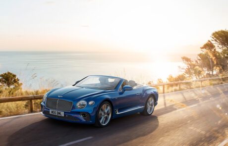 A sleek blue Bentley Azure convertible car is driving on a coastal road at sunset. The sea and distant shoreline are visible in the background, with the sun casting a golden glow over the scenery. Tall grass and trees partially line the road, as if crafted by CGI Studio London.