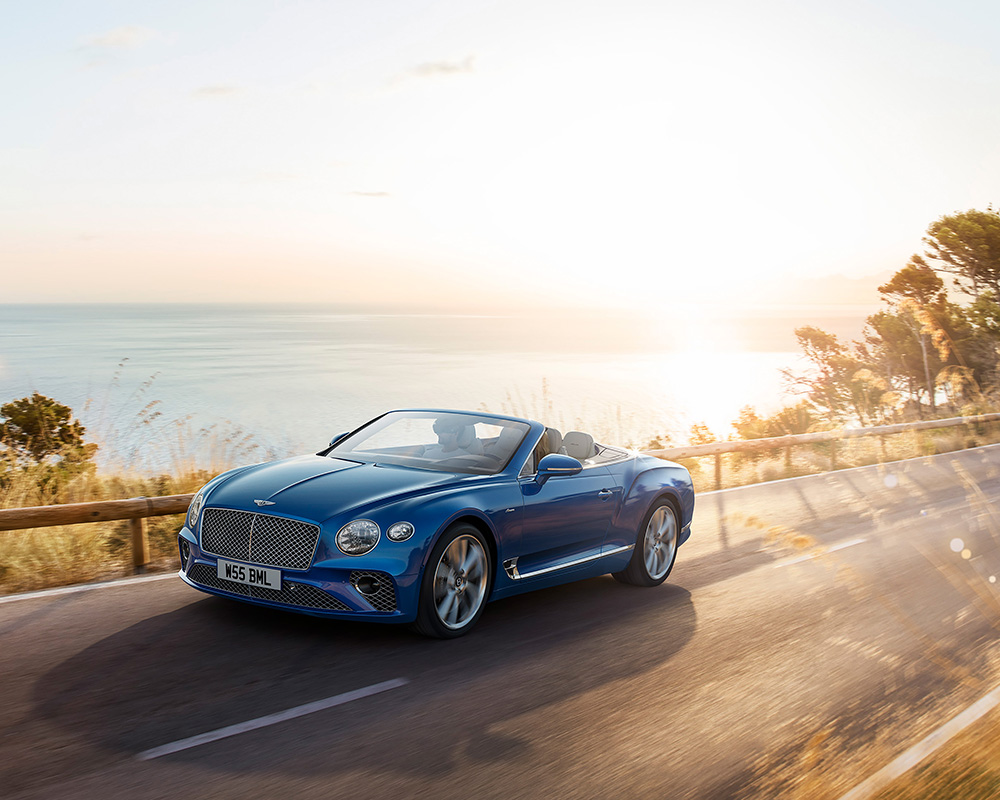 A sleek blue Bentley Azure convertible car is driving on a coastal road at sunset. The sea and distant shoreline are visible in the background, with the sun casting a golden glow over the scenery. Tall grass and trees partially line the road, as if crafted by CGI Studio London.