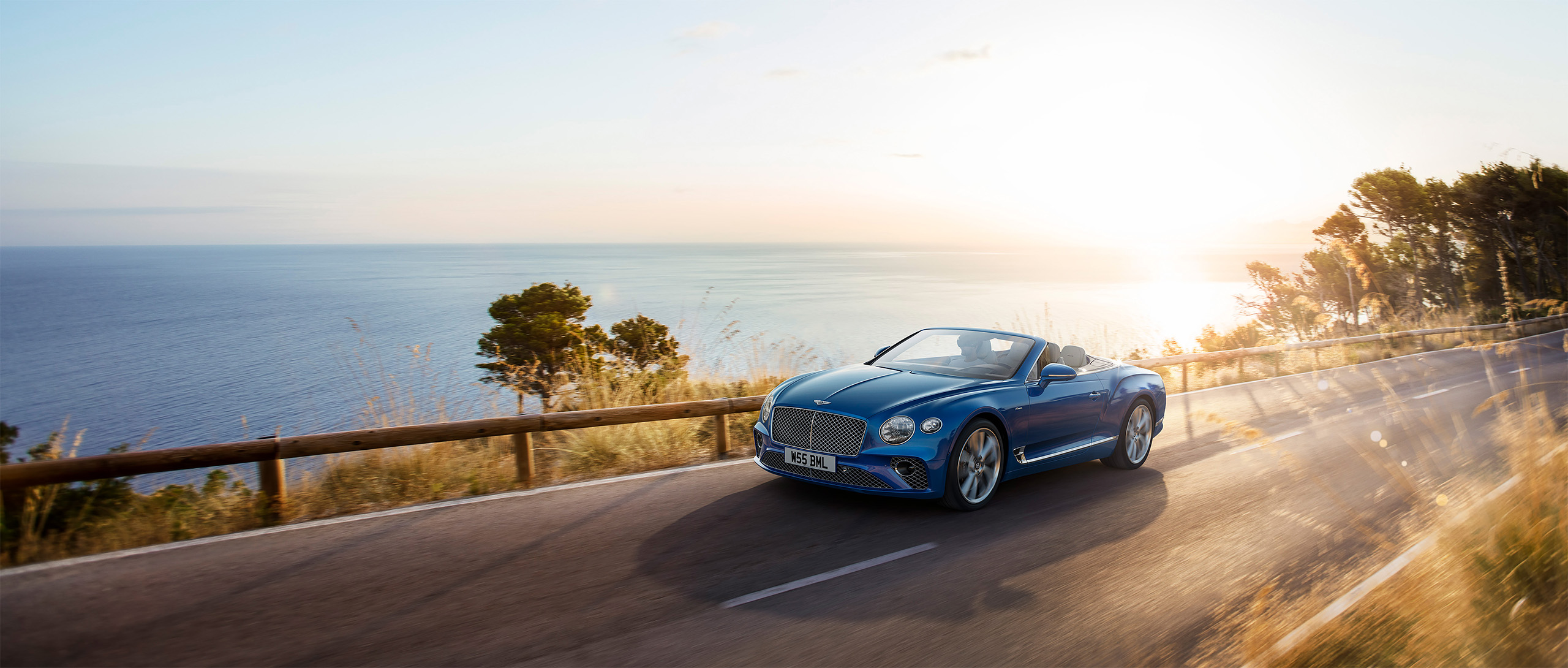 A blue Bentley Azure convertible drives along a coastal road with the ocean and a bright setting sun in the background. The scene, rendered by CGI Studio London, is bordered by trees and grasses, creating a picturesque and serene ambiance.