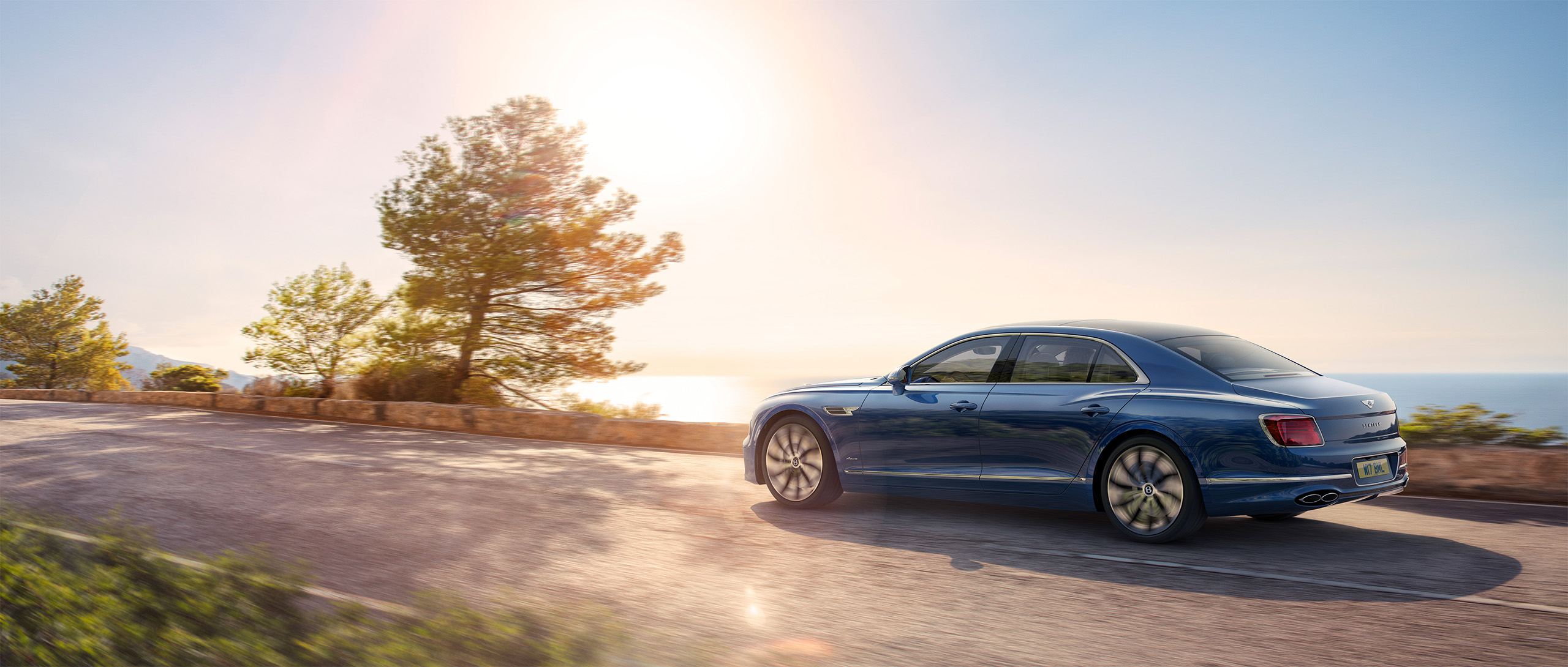 A sleek blue Bentley Azure sedan drives along a scenic coastal road bathed in sunlight, the ocean and trees in the background. The sun shines brightly, casting a warm glow over the tranquil setting. This stunning scene was masterfully rendered by CGI Studio London.