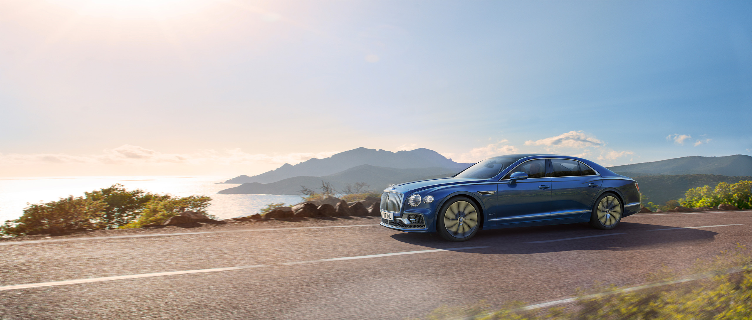 A luxurious Bentley Azure sedan drives along a scenic coastal road with a backdrop of mountains, the sun shining brightly in a clear sky. The ocean is visible to the left, reflecting the sunlight. The landscape is lush and picturesque, as if crafted by a CGI studio in London.